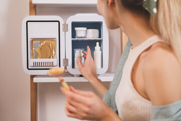 Woman with face roller taking cosmetic product from mini fridge indoors, closeup