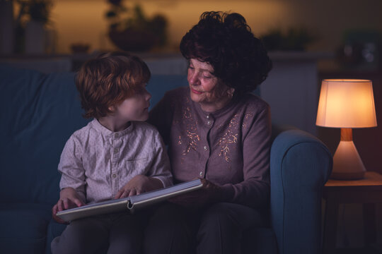 Kind Grandma Reading A Story From A Book To Her Grandson, Sitting On The Couch In A Living Room In The Evening