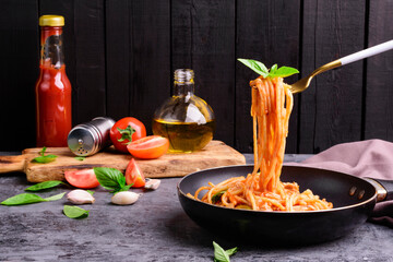 A fork is scooping up classic Italian spaghetti in a black pan with tomato sauce and basil. Freshly prepared food on a wooden table on a dark wooden wall background