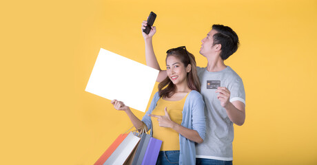Asian couple holding blank billboard with credit card and shopping bags and smartphone isolated on yellow background