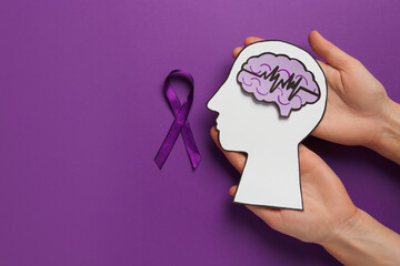 Top view of woman holding paper human head cutout with brain near purple ribbon on color background, space for text. Epilepsy awareness