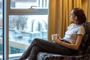 woman sits in a chair by the window of a high floor and looks at the city view