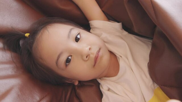 Face focusing 4K clip of adorable asian girl kid laying down looking up and playing with camera in joyfulness, happiness and relax emotion shows comfortable expression at home with family.