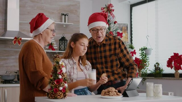 Happy family watching online childhood xmas video using tablet computer celebrating christmas holidays in decorated kitchen. Children eating baked cookies drinking milk enjoying winter season