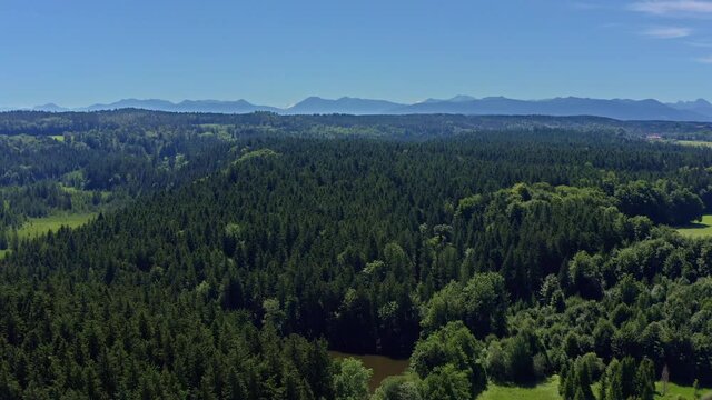 Wide green forest - smooth drone flight backwards in mountain range with a little pond called Moosbach in the foreground - idyllic landscape in southern bavaria, germany.