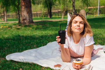 Fototapeta premium Young romantic woman laying on the grass in park with open book, cake and cup of coffee to go