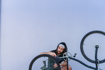 young latino hispanic man with long hair repairing his bicycle. grey wall background, isolated, copy space