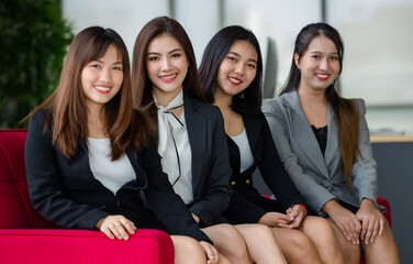 Group of four attractive Asian female office colleagues in formal business suits sitting on red couch in office smiling at camera. Concept for modern office working