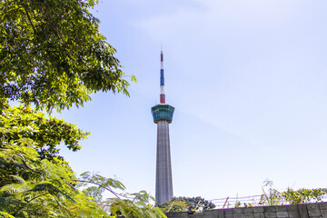 Obraz premium Lighthouse and traffic control tower cargo ship sailing as national flag picture green tree with outside concrete fence and barbed wire blur pictures front blue sky clouds background, symbol landmark 