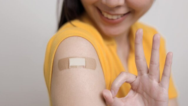 Close up young beautiful asian woman in yellow shirt getting a vaccine protection the coronavirus. Smiling happy female showing arm with bandage after receiving vaccination.Hand Showing  ok,thums up