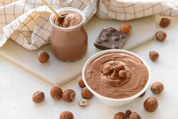 Jar and bowl with tasty chocolate paste and hazelnuts on light background