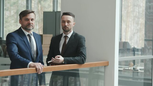 Medium PAN slowmo portrait of two successful male lawyers in suits and ties looking at camera standing by railing in contemporary office