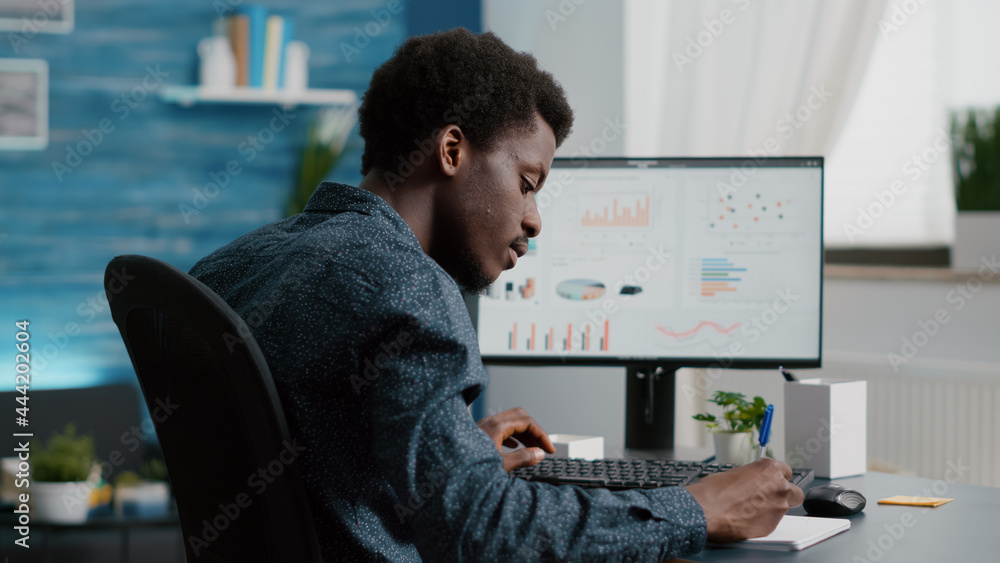 Poster selective focus on black american guy working from home, taking notes on notepad while remote workin
