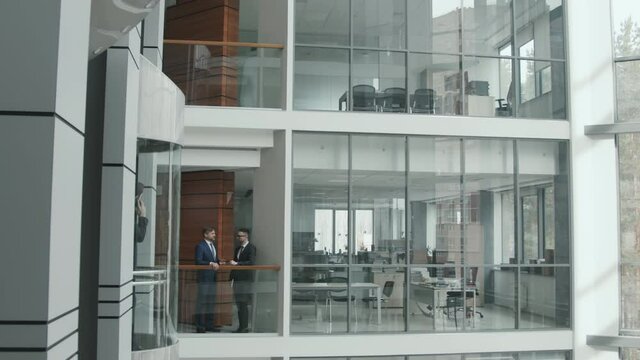 Wide Slowmo Shot Of Two Male Lawyers In Suits Working In Big Firm Having Conversation Standing By Railing In Modern Office