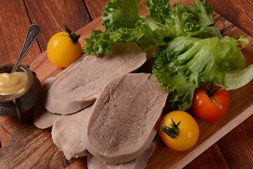 Sliced Beef Tongue Slices on a platter with lettuce leaves, cherry tomatoes and Dijon mustard on a wooden background