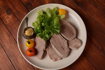 Sliced Beef Tongue Slices on a platter with lettuce leaves, cherry tomatoes and Dijon mustard on a wooden background