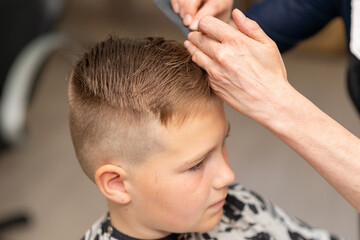 Cute european boy getting hairstyle, hairdresser makes a hairstyle for a boy.Side view.