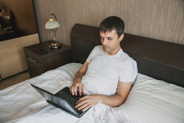 A middle-aged man is sitting in bed in his bedroom and working on a laptop