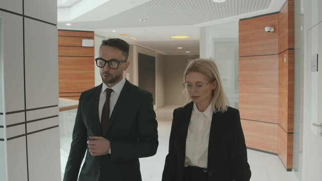 Medium tracking shot of couple of lawyers or business partners in formalwear talking while walking through modern office
