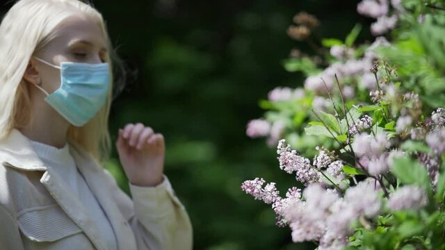 A young blonde woman walks up to the lilac bushes, takes off her medical mask and enjoys the scent of flowers. Happy woman without mask in the park. Close-up, slow motion, HD.