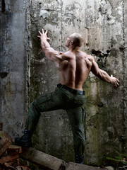 Muscular bodybuilder with a naked torso in camouflage military pants and boots stands among the old concrete, moldy walls. Rear view. Ruins, fragments of slabs and bricks. Light penetrating from above