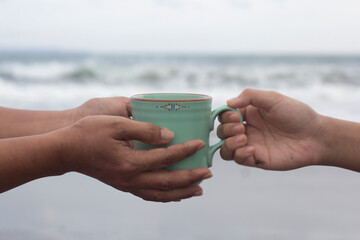 Hands of two people holding a cup of coffee or tea on beach background. Take and give or giving and receive concepts. Kindness concept. - obrazy, fototapety, plakaty