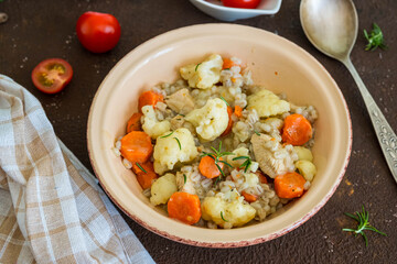 Hearty stew with chicken, barley, carrots and cauliflower in a ceramic bowl on a brown concrete background.