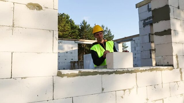 Construction worker at construction site puts the wall of a house made of aerated concrete blocks. Protective clothing - hardhat and a vest, the construction process