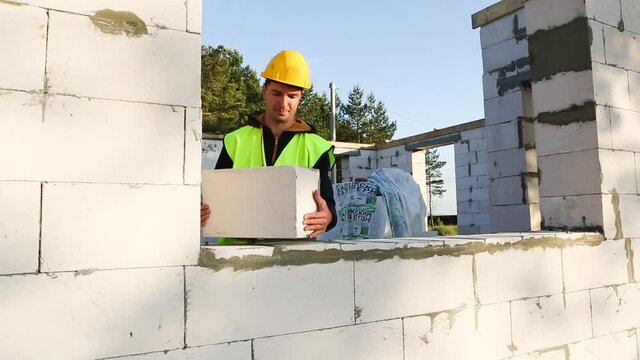Construction worker at construction site puts the wall of a house made of aerated concrete blocks. Protective clothing - hardhat and a vest, the construction process