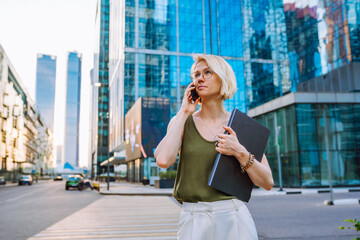Fototapeta premium Young business woman standing with laptop among the skyscrapers and speaks on the phone.