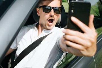 Portrait of a surprised young man with an open mouth filming an event on a mobile phone while driving a car. Selective focus on the shocked face of the driver.