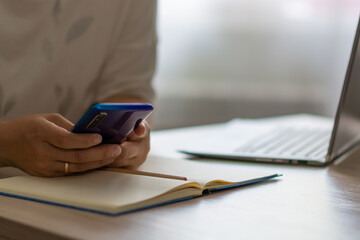 Female hands work in the phone near the laptop and notepad. Married woman working at home office. Horizontal view.