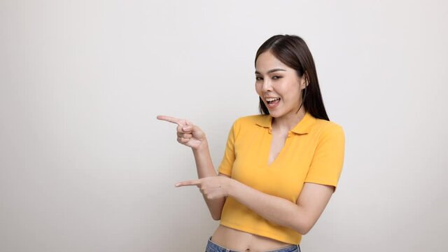 Excited asian woman pointing the finger to blank space for advertising text on isolated background. Joyful teenage girl in yellow shirt standing in white room looking at empty space.