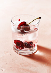 Glass of Summer cocktail with gin and cold tonic, decorated with halved cherries, few red peppercorns and with ice cubes on pink background. Sunny day shadows.