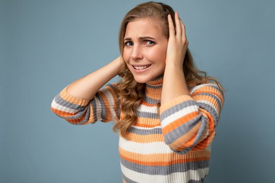 Photo of young emotional angry pretty nice blonde curly woman with sincere emotions wearing casual striped pullover isolated on blue background with free space and covering ears trying not to hear