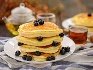 Sweet food. Stack of delicious pancakes with blueberries and honey, tea, plaid fabric in white plate on blur background.