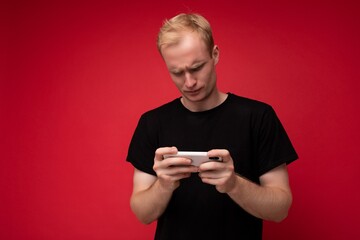 Photo of handsome concentrated upset dissatisfied young blonde man isolated over red background wall wearing black t-shirt holding and using mobile phone watching something via smartphone looking down