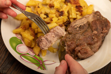 Someone is cutting a piece of juicy roast meat on a plate of fried potatoes with a knife and fork. Close-up Selective focus.