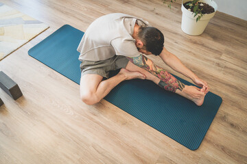 Top view of tattooed young man stretching on yoga mat reaching for toes with hands