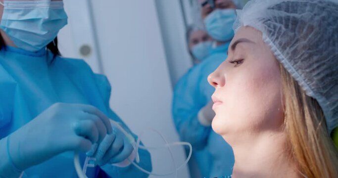 A female patient is sitting in the dentist's chair while a female medical worker in a disposable mask and gown is inserting into her nose an oxygen nasal tube.