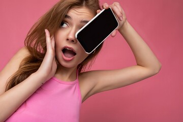 Naklejka premium Closeup portrait photo of sexy surprised beautiful positive young blonde woman wearing pink top poising isolated on pink background with empty space holding in hand and showing mobile phone with empty
