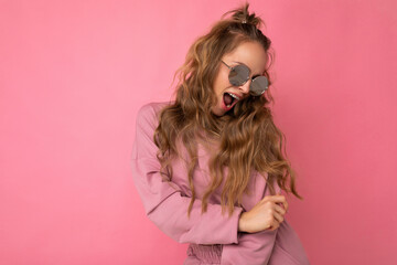 Shot of fascinating happy young dark blonde curly woman isolated over pink background wall wearing casual pink sport clothes and stylish sunglasses looking to the side and having fun