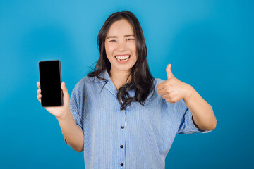 Portrait of happy Asian woman wears a striped cyan shirt her thumb up and holding mobile phone or smartphone isolated on blue background, Mock up copy space