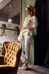 photo of a beautiful dark-skinned girl standing around the sofa in a white blouse with yellow flowers and white pants and looking down