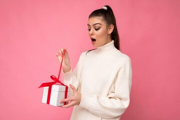 Photo shot of pretty positive surprised young brunet woman isolated over colourful background wall wearing trendy outfit look holding gift box and looking at present box with red ribbon