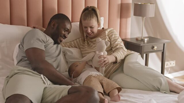 Medium long of happy diverse parents lying in bed, playing with their Mixed-Race toddler and stuffed bear toy, smiling