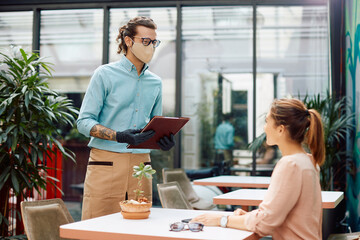 Happy waiter with face mask holding menu and talking to female guest in a restaurant.