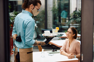 Waiter with face mask and gloves serving female customer while working at reopened cafe.