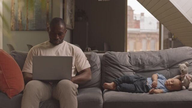 Medium long of young African man sitting on couch, typing on portable computer on his lap, little adorable child sleeping nearby