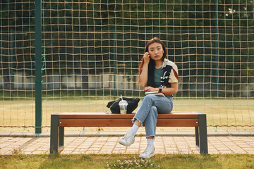 Sits against sport field. Young asian woman is outdoors at daytime